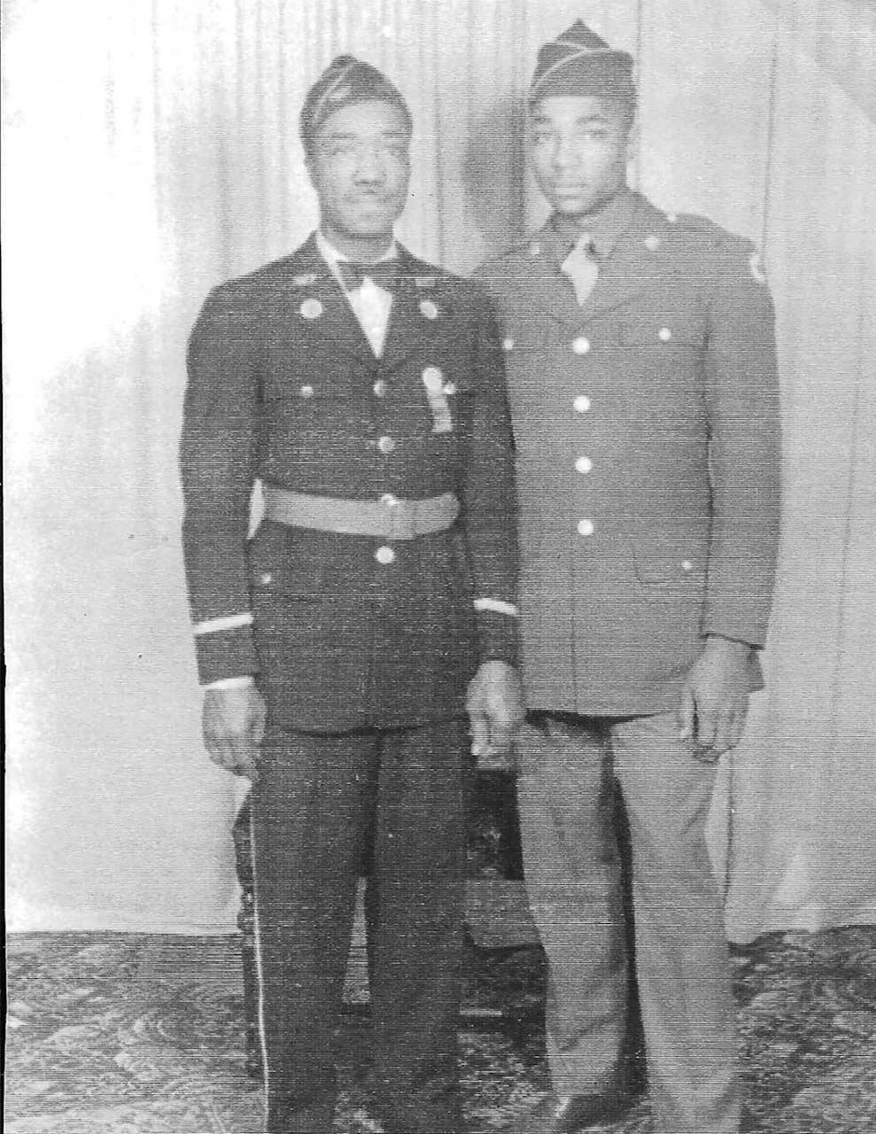 Faded black-and-white full-length portrait of two uniformed men against a curtain.