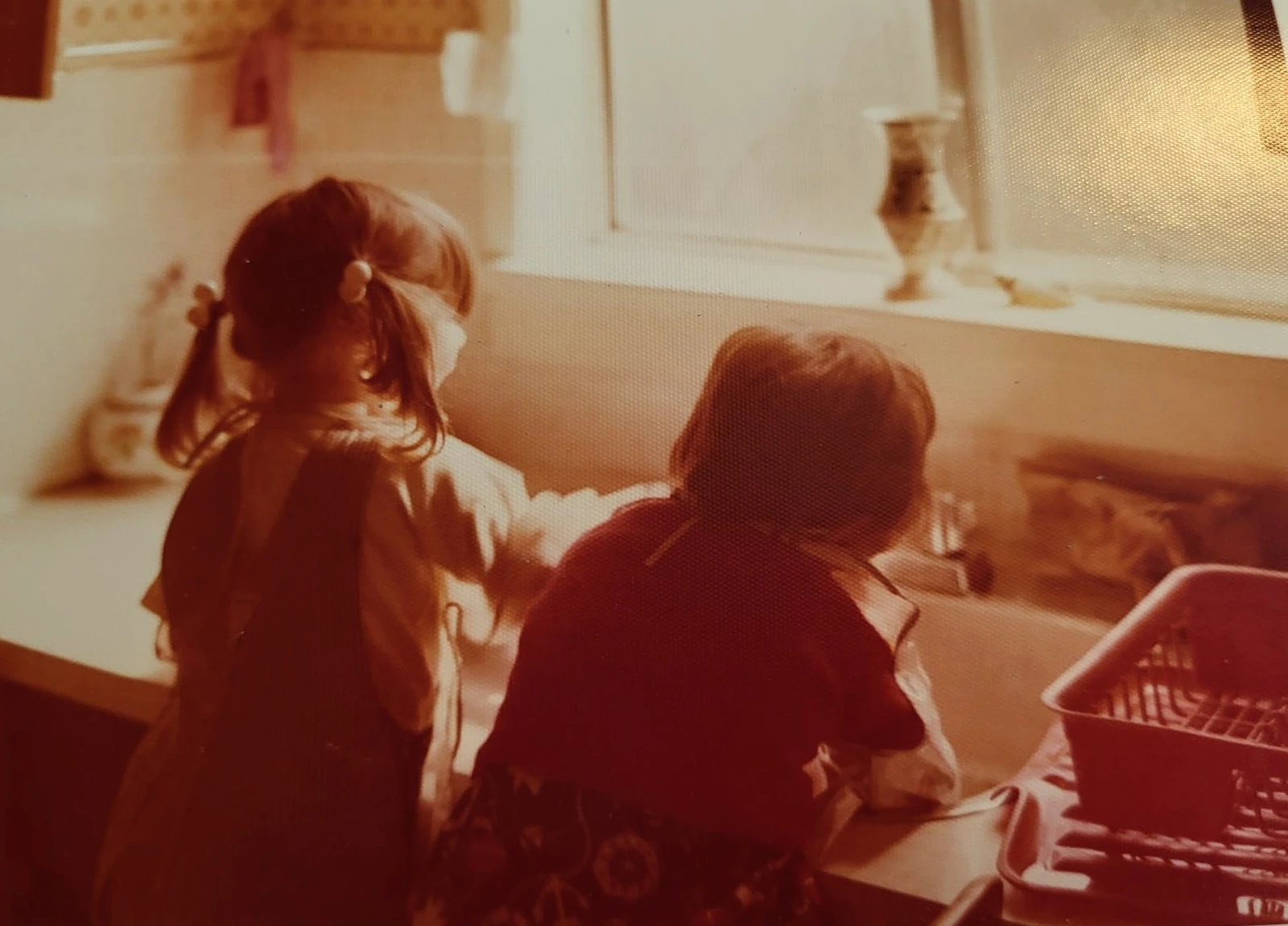 Warm orange-red photograph of two children at a kitchen sink, with soft detail and visible print texture.